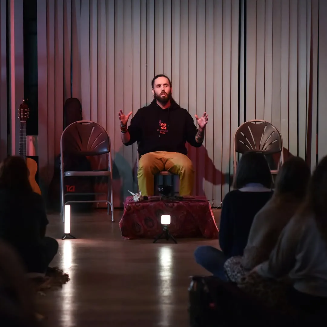 UJJAY seated on a low stage in a wood-panelled room, hands raised mid-sentence, speaking to an audience sitting on the floor around him.