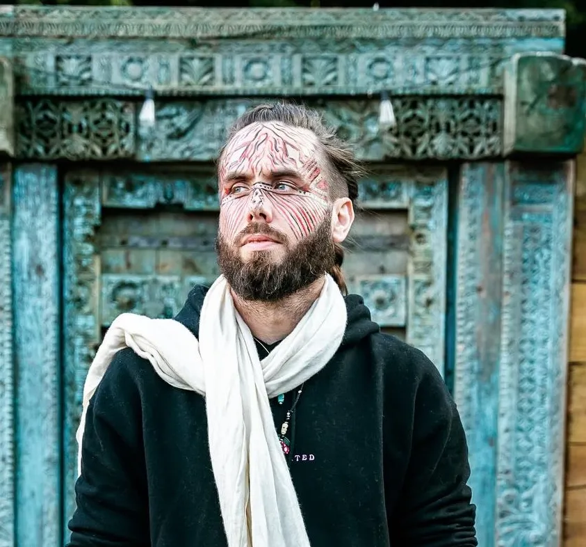 UJJAY, wearing fine red and white linework across his face, a black hooded top and a cream scarf, standing before a weathered turquoise carved doorway.